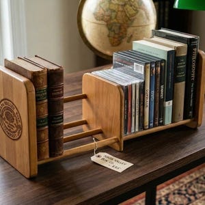 May include: An adjustable wooden book rack with two vertical end panels, one featuring a circular Napa Valley Box Company logo. The rack has three horizontal connecting rods and holds two vintage books on the left and several books and CDs on the right.
