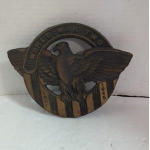 May include: A circular bronze-toned metal plaque featuring a central eagle with outstretched wings. The text WORLD WAR TWO is embossed along the top arch, while the years 1941 and 1945 are vertically inscribed on the lower striped base section.
