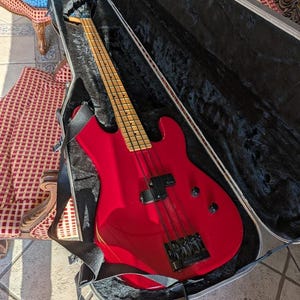 A bright red electric bass guitar in an open black case. The bass has a maple neck and fretboard, black hardware, and a black strap. The case is open, revealing the instrument's body and neck. Available at SecondhandSandies