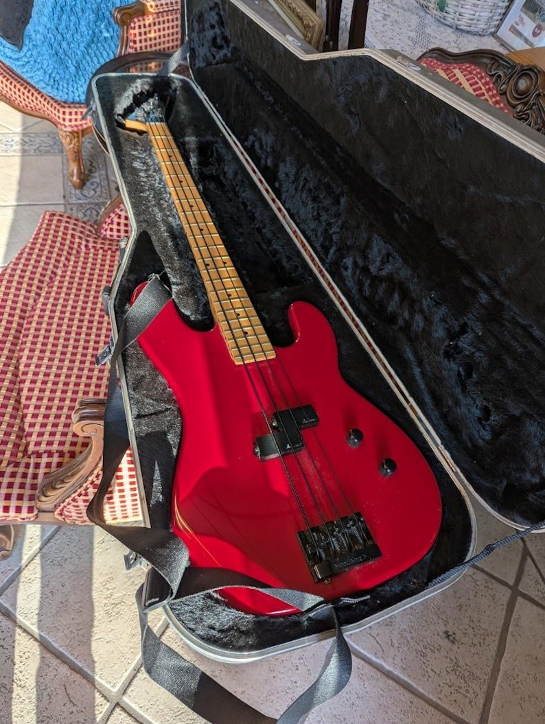 A bright red electric bass guitar in an open black case. The bass has a maple neck and fretboard, black hardware, and a black strap. The case is open, revealing the instrument's body and neck. Available at SecondhandSandies