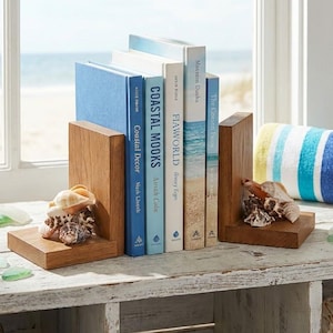 May include: Wooden bookends with seashells supporting a collection of books with blue and white covers. A jar filled with sand, seashells, and a starfish sits nearby. A striped towel and sea glass complete the coastal decor.