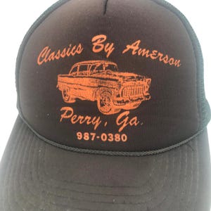 May include: A black trucker hat with an orange graphic of a classic car and the text "Classics By Amerson, Perry, Ga, 987-0380". The hat has a mesh back and a curved brim.
