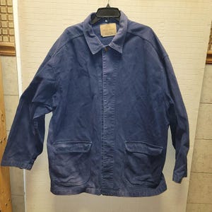 May include: A blue work jacket with a collared neckline and two large front pockets. The jacket has a button-up closure and a small label near the collar. The jacket is hanging on a black hanger.