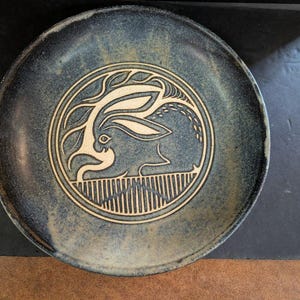 May include: A round ceramic plate with a blue and tan glaze. The center features a cream-colored rabbit design within a circular border. The rabbit is depicted in a stylized, artful manner, with long ears and a detailed outline.