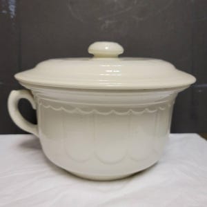 May include: Off-white ceramic chamber pot with a matching lid and handle. The pot features a scalloped design around the rim and vertical grooves. The lid has a small, round knob for easy removal.