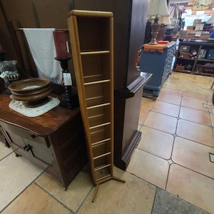 May include: A tall, narrow wooden shelf unit with multiple horizontal shelves. The unit is made of light-colored wood and has a simple, minimalist design. It stands on two angled legs and is suitable for displaying books or decorative items.