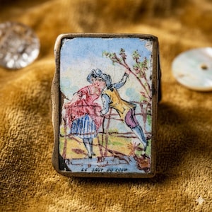 May include: Antique rectangular trinket box with a painted scene of two figures near a tree, with the text "LE SAUT DU LOUP" at the bottom. The box has a gold-colored metal frame and is set against a gold velvet surface.