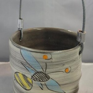 May include: A cylindrical ceramic bucket with a grey and white swirled finish, featuring a hand-drawn bee illustration with blue wings and yellow stripes. The text "Bee the ch" is written in black script. It has a metal wire handle with a ceramic grip.