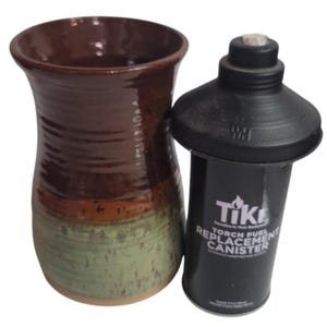 May include: A brown and green glazed ceramic vase with a flared rim next to a black Tiki brand torch fuel replacement canister. The canister features white text reading Tiki, Torch Fuel Replacement Canister, and a capacity of 12 fl oz.