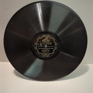 May include: A black circular vinyl phonograph record featuring a central black label with gold text and graphics. The label displays the Victor brand logo, the title The Sweetest Story Ever Told, and the artist name Neapolitan Trio, number 20278-A.