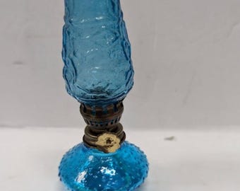 May include: A miniature oil lamp featuring a bright blue glass base with a textured hobnail pattern and a matching blue glass chimney with a wavy, embossed surface. The burner assembly is made of aged brass and includes a small, circular adjustment knob.