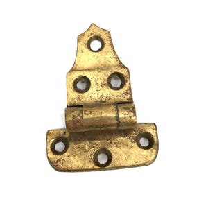 May include: A vintage brass hinge with a decorative top and a rectangular base. The hinge has a tarnished, aged appearance, with visible wear and tear. It features multiple screw holes for mounting and a central barrel for movement. The overall color is a warm, antique gold.