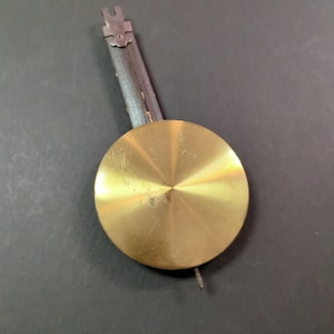 May include: A brass clock pendulum with a circular weight and a dark wooden rod. The pendulum has a small, decorative top piece. The brass weight has a brushed finish and a small, pointed tip.