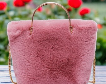 Könnte beinhalten: Eine rechteckige, rosa Handtasche mit flauschiger Kunstfellstruktur. Die Tasche hat einen goldfarbenen Griff und einen goldfarbenen Kettenriemen. Der Hintergrund ist verschwommen, mit roten Blumen.