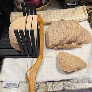 May include: A black bread slicer with a wooden handle, used to slice a loaf of brown bread. The bread is sliced into even pieces and placed on a white kitchen towel.