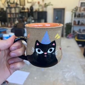 May include: A ceramic mug with a black cat wearing a blue party hat. The mug has a tan background with orange rim and colorful accents.