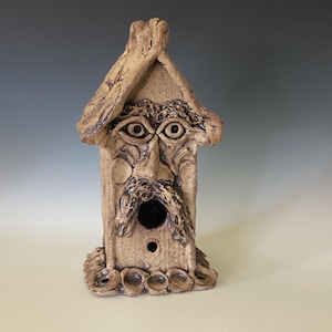 May include: A whimsical, handmade birdhouse crafted from tan-colored ceramic. The birdhouse features a stylized face with large eyes, a mustache, and a beard. The roof has a textured, bark-like appearance, and the base has decorative elements.