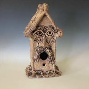Könnte beinhalten: Ein skurriles, handgefertigtes Vogelhaus mit einem Gesicht, das in die Vorderseite geschnitzt ist. Das Vogelhaus besteht aus hellbrauner Keramik und hat einen detaillierten Schnurrbart, Augen und eine kleine runde Öffnung für Vögel. Das Dach hat eine strukturierte Optik.