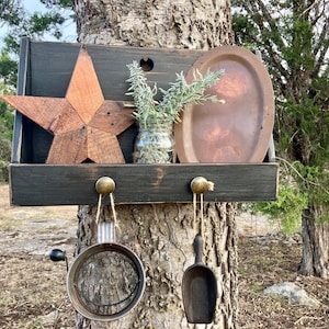 May include: A rustic wooden shelf with a star, a metal plate, and a small glass jar with greenery. Below, two metal kitchen tools hang from brass knobs. The shelf is mounted on a tree trunk, creating a charming, country-style display.