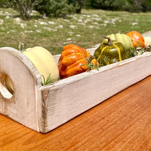 May include: A rustic, rectangular wooden trough filled with a variety of small pumpkins in shades of orange, yellow, and green. The trough has a distressed white finish and is decorated with sprigs of greenery.