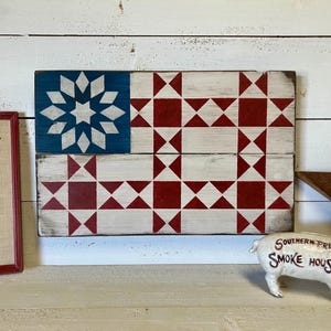 May include: A wooden American flag with a blue starburst design and red and white geometric patterns. The flag is displayed on a white wooden surface. A white pig-shaped bank with "Southern Pride Smoke House" text is in the foreground.