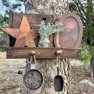 May include: A rustic wooden shelf with a star, a jar of greenery, and a metal plate. Below, two hooks hold a strainer and a scoop. The shelf is mounted on a tree trunk, creating a charming, country-style display.
