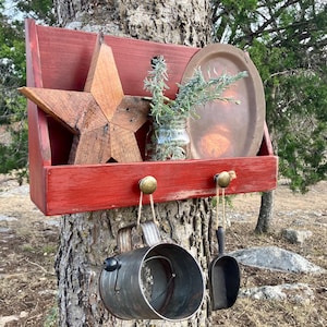 May include: A rustic, red wooden shelf with a star, a metal plate, and a small glass jar with greenery. Below, a metal bucket and scoop hang from brass knobs, creating a charming, country-style display.
