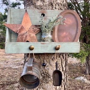 May include: A rustic, wooden shelf painted in a distressed light green, mounted on a tree. It features a wooden star, a copper tray, a glass jar with greenery, and hanging metal and wooden accessories. The shelf is decorated with a vintage aesthetic.