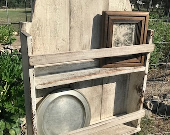 May include: A whitewashed wooden shelf with two shelves and a rounded top. The shelf has a small hole in the top and is attached to a wire fence. A silver plate and a framed picture are on the shelves.