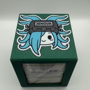 May include: A green card storage box with a cartoon graphic of a character with blue hair and the text "JENOVA" on the lid. The box is cube-shaped and appears to be made of plastic.