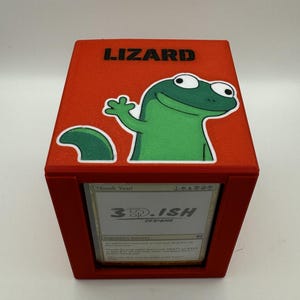 May include: Red card storage box with a green cartoon lizard graphic and the word "LIZARD" in black. The box has a card visible with the text "3D-ISH DESIGNS".