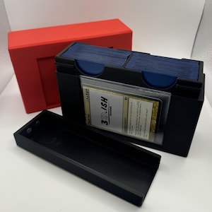 May include: Black card storage box with a red lid. The box has two compartments for cards and a separate tray. A card in a protective sleeve is visible, with the text "3D-ISH DESIGN".