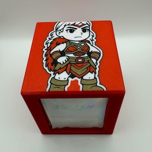 May include: A red rectangular box with a cartoon illustration of a female warrior. The warrior has blonde hair, a stern expression, and is wearing a red and gold outfit. The box has a red frame and the text "3RD.ISH" is visible.