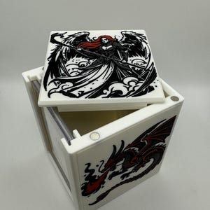 May include: White box with a hinged lid, featuring black and red fantasy art. The lid displays a woman with red hair and wings, holding a staff. The side of the box shows a red and black dragon breathing fire.