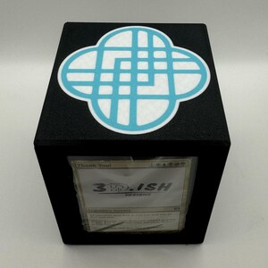 May include: Black cube-shaped card holder with a light blue and white design on top. The front of the holder displays a card with the text "3D.ISH DESIGNS" and "Thank You!" printed on it. The holder is designed to store cards.
