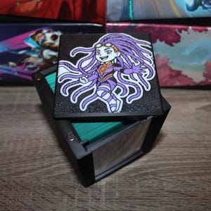 May include: A black and green card deck box with a purple and white illustration of a cartoon character with long purple hair and a white shirt. The character is wearing a purple and white outfit and has a white headband.