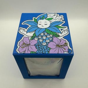 May include: A blue box with a floral design featuring a sleeping face with blue and white hair, blue and purple flowers. The box has the text "3D.ISH" on the front.