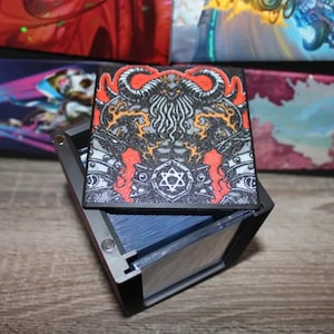 May include: A black card storage box with a detailed, colorful design on the lid. The artwork features a central figure with horns, surrounded by intricate patterns in red, orange, and white. The box is open, revealing a stack of blue cards.