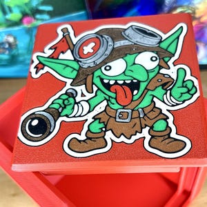 May include: A red box with a cartoon goblin illustration. The goblin is green with a brown hat, goggles, and a brown outfit. It holds a telescope and gives a thumbs-up. The goblin has a red tongue sticking out.