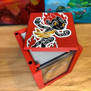 May include: Red card storage box with a cartoon character design on the lid. The box is open, revealing a stack of cards and a clear plastic divider. The character is black with gold accents and a flame-like head.