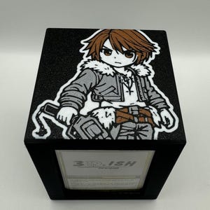 May include: A black, cube-shaped box with a cartoon illustration on top. The illustration features a character with brown hair, a gray jacket, and a white outline. The box has a framed opening with the text "3D-ISH DESIGNS".