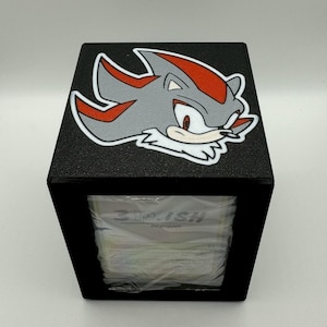 May include: Black square tissue box featuring a gray and red character design. The tissue box has a textured surface and a rectangular opening. The text "3DISH DESIGNS" is visible on the tissue packaging.