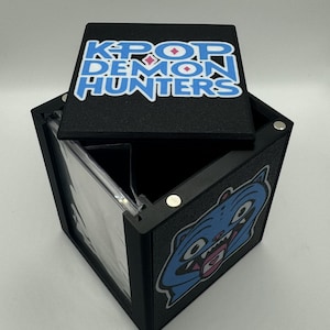 May include: Black box with the text "K-POP DEMON HUNTERS" on the lid. The box has a blue and pink graphic on the lid and a blue cartoon cat graphic on the side. The box is open, revealing a clear plastic insert.