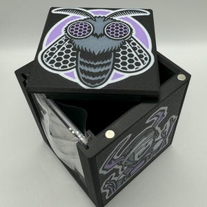 May include: Black, square box with a hinged lid. The lid features a moth illustration in gray, purple, and white. The box is open, revealing a clear plastic interior. The box has a magnetic closure.