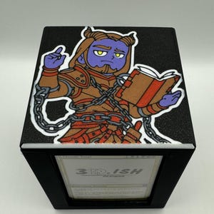 May include: Black card storage box with a cartoon illustration of a purple-skinned character reading a book. The box has a black exterior and a white interior. The text "3D-ISH DESIGNS" is visible on the front.