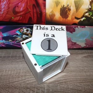 May include: White card deck box with a white lid that says "This Deck is a 1". The lid is open and the box is filled with teal colored cards.
