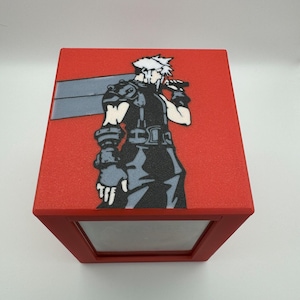 May include: Red box featuring a graphic of a character with spiky white hair, a large sword, and black clothing. The character is set against a red background. The box has a clear window on the front.