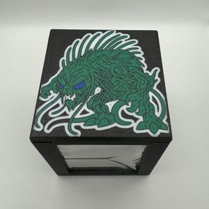 May include: Black box with a green monster design on the top. The monster has blue eyes and a white outline. The box has a clear front, and the design is on a textured black surface.