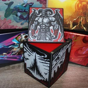 May include: A black and red box with a detailed illustration of a muscular demon with horns and wings. The box is open, revealing a red interior. The exterior features a black and white castle design. Other boxes with fantasy art are in the background.