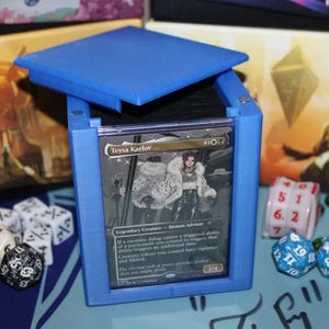 May include: A blue deck box with a clear plastic window showing a Magic: The Gathering card featuring Teysa Karlov, a legendary creature. The box is open and the lid is propped up. There are dice scattered around the box.